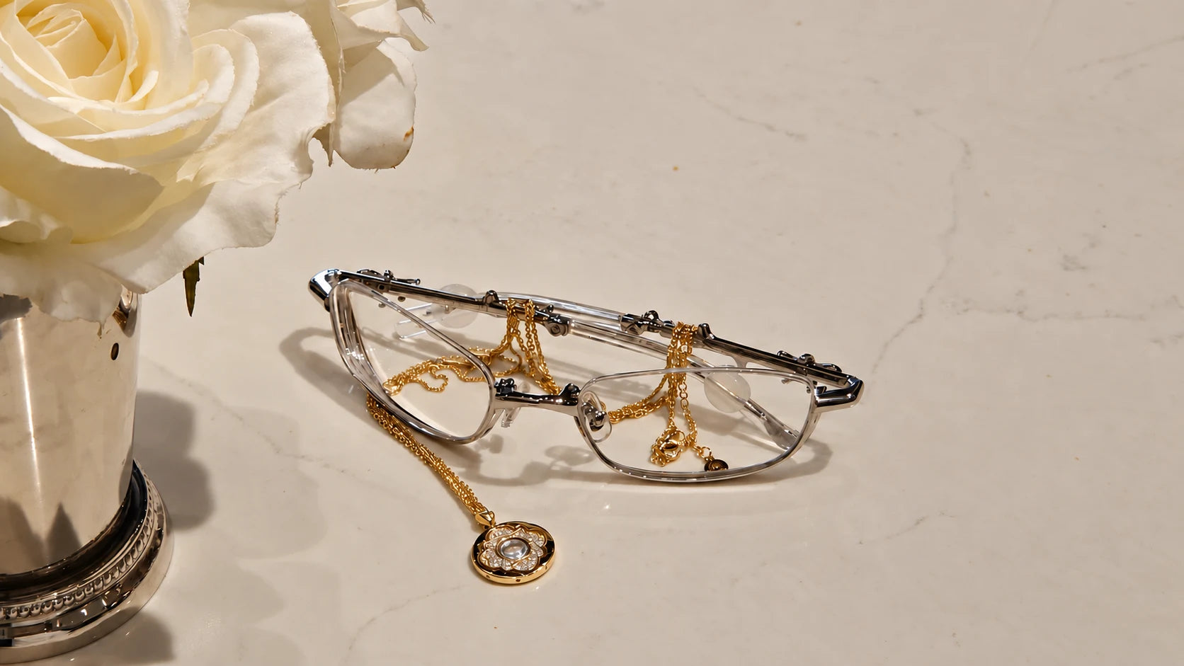 The gold Blessed Sand Necklace draped over glasses beside white roses